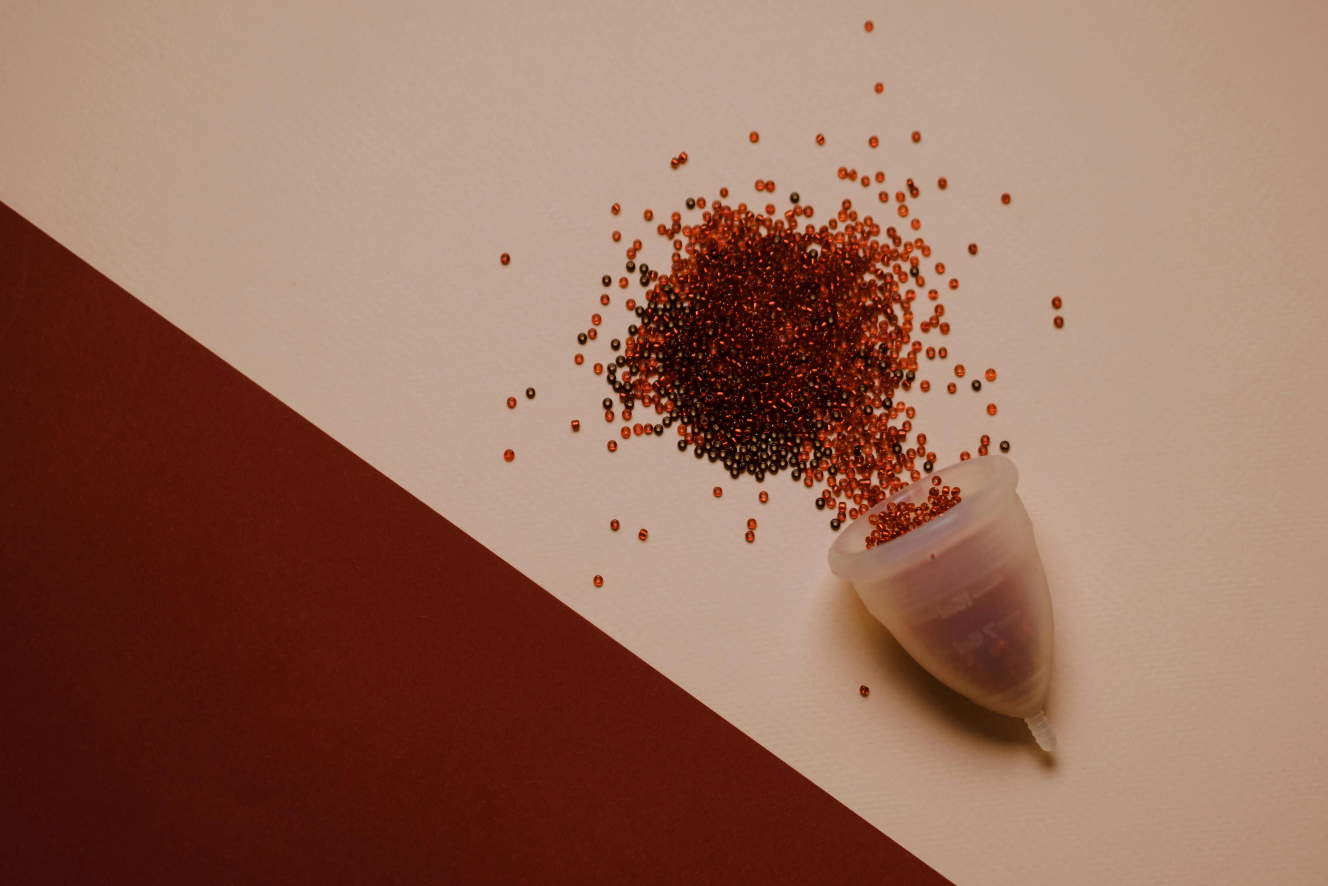 Visual Representation of Menstruation: Menstrual Cup and Red Seeds Menstrual cup surrounded by scattered red seeds symbolizing menstrual blood